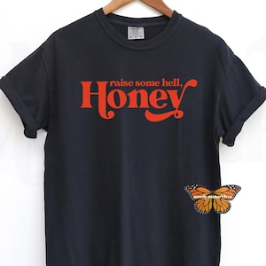以下が含まれることがあります： 「raise some hell, Honey」という赤い文字が書かれた黒いTシャツ。