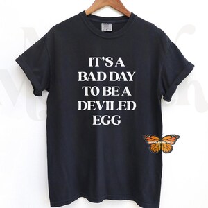Puede incluir: Camiseta negra con texto blanco que dice "Es un mal día para ser un huevo diabólico."
