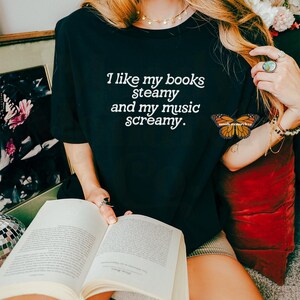 May include: A woman wearing a black t-shirt with the text "I like my books steamy and my music screamy." She is holding a book and sitting on a red cushion.