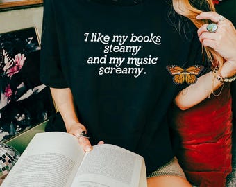 May include: A woman wearing a black t-shirt with the text "I like my books steamy and my music screamy." She is holding a book and sitting on a red cushion.