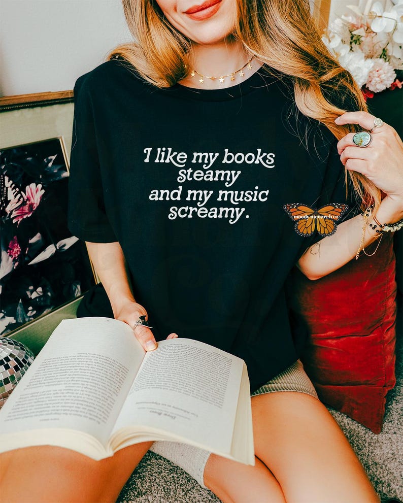 May include: A woman wearing a black t-shirt with the text "I like my books steamy and my music screamy." She is holding a book and sitting on a red cushion.