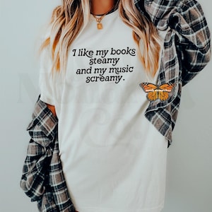 May include: White t-shirt with the text "I like my books steamy and my music screamy." printed in black. A butterfly sticker is on the bottom right corner of the shirt.