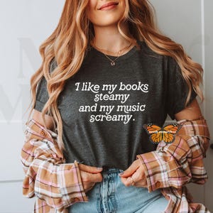 May include: A dark gray t-shirt with the text "I like my books steamy and my music screamy." printed in white. The shirt is being worn by a person with long blonde hair and a plaid shirt.