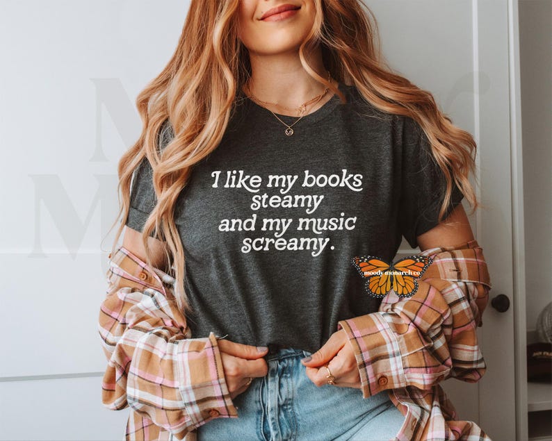 May include: A dark gray t-shirt with the text "I like my books steamy and my music screamy." printed in white. The shirt is being worn by a person with long blonde hair and a plaid shirt.