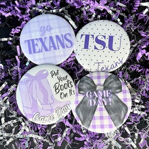 May include: Four round buttons with purple and white designs. One button says "go TEXANS" on a plaid background. Another says "TSU Texans" with polka dots. A third has a boot graphic and says "Put Your Boots On Its Game Day". The last says "GAME DAY!" with a bow.