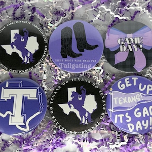 May include: A collection of six round buttons with purple and white designs. The buttons feature text and graphics related to a university, including a map of Texas, cowboy boots, and the words "Game Day!"