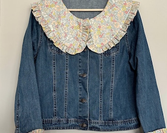 May include: A blue denim jacket with a large, ruffled collar made of a floral fabric. The collar has a white background with pink, yellow, and blue flowers.