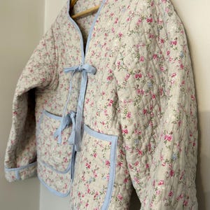 May include: A quilted jacket with a floral pattern of pink and blue flowers on a cream background. The jacket has light blue trim, two front pockets, and a light blue tie closure. The jacket is hanging on a wooden hanger.