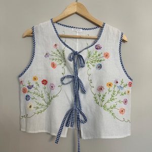 May include: White sleeveless top with colourful embroidered flowers and green leaves. The top has a blue and white gingham trim and a matching tie closure. The floral embroidery includes red, yellow, orange, and purple flowers.