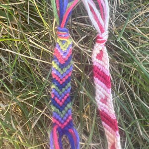May include: Two handmade friendship bracelets. One features a chevron pattern in shades of purple, blue, green, and pink. The other is a striped design in red, pink, and white. Both are made of colorful threads and are displayed on a bed of grass.