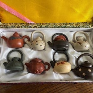 以下が含まれることがあります： 装飾的な箱に入った8つのミニチュアティーポットのセット。ティーポットは、茶色、黒、ベージュなど、さまざまな色があり、さまざまなデザインが施されています。箱は赤と金色の外観と白い内装です。