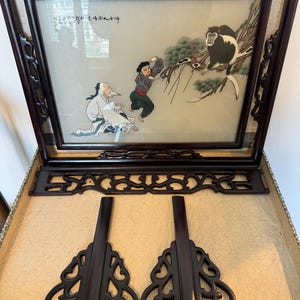 May include: An antique framed artwork with a detailed illustration of figures and a monkey. The frame is dark brown with ornate carvings. The artwork is accompanied by two decorative stands and a decorative top piece, all in matching dark brown wood.