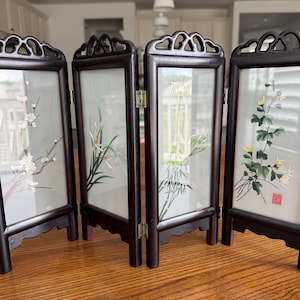 May include: A four-panel folding screen with dark wood frames. Each panel features a different floral design in shades of white, green, and yellow. The screen is designed to stand upright on a table or other flat surface.