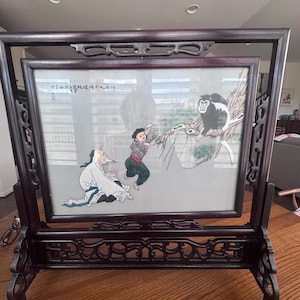 May include: A framed Asian artwork featuring a painted scene with figures and a monkey. The dark wood frame has intricate carved details. The artwork depicts a man in white robes, a person in red and black clothing, and a monkey in a tree.