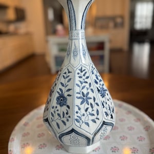 Peut inclure: Un vase en céramique blanc et bleu avec un motif floral. Le vase a une forme hexagonale et un dessus évasé. Il repose sur un plateau décoratif avec un motif floral. Le vase est un objet décoratif.