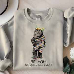 Może przedstawiać: Jasnoszara bluza z nadrukiem postaci podobnej do Groota obejmującej pluszowego misia z puzzli. Postać ma na głowie słoneczniki. Tekst "BE YOU! THE WORLD WILL ADJUST" jest wydrukowany poniżej.