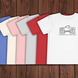 Puede incluir: Cinco camisetas de diferentes colores, incluyendo azul, crema, rosa, gris y rojo, est&aacute;n alineadas en una superficie de madera. La camiseta de la derecha tiene una ilustraci&oacute;n en blanco y negro de una camioneta con neum&aacute;ticos grandes.