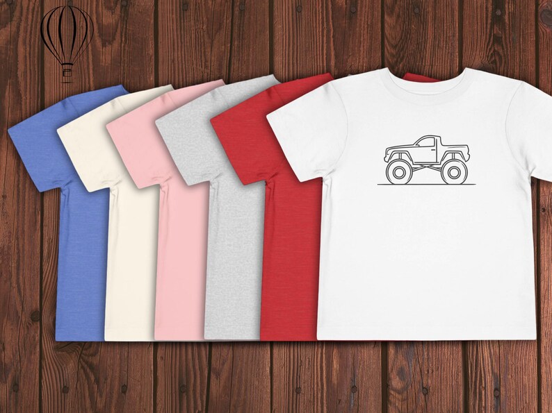 Puede incluir: Cinco camisetas de diferentes colores, incluyendo azul, crema, rosa, gris y rojo, est&aacute;n alineadas en una superficie de madera. La camiseta de la derecha tiene una ilustraci&oacute;n en blanco y negro de una camioneta con neum&aacute;ticos grandes.