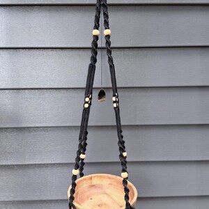 May include: A black macrame plant hanger with a wooden bowl. The hanger is adorned with wooden beads and a decorative tassel. The plant holder is suspended from a metal ring.