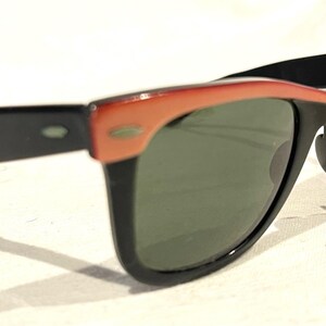May include: Sunglasses with a black frame and dark green lenses, featuring a coral-coloured upper frame. The design is retro, with a small logo on the lens. The arms are black, and the style is suitable for all genders.