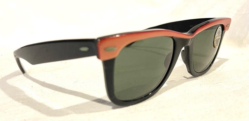 May include: Sunglasses with a black frame and dark green lenses, featuring a coral-coloured upper frame. The design is retro, with a small logo on the lens. The arms are black, and the style is suitable for all genders.