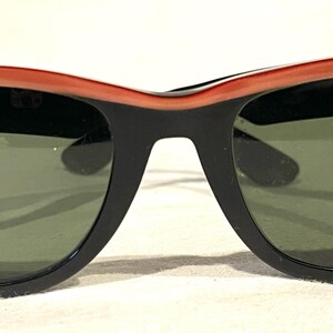 May include: Black and coral sunglasses with green lenses. The sunglasses feature a classic design with a thick black frame and a coral-coloured top frame. A small, round sticker is on the right lens.