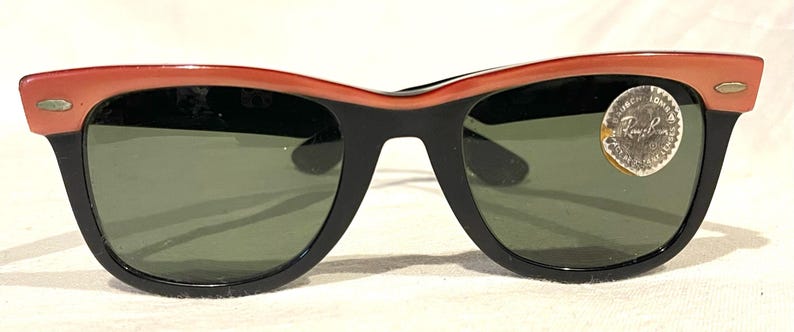 May include: Black and coral sunglasses with green lenses. The sunglasses feature a classic design with a thick black frame and a coral-coloured top frame. A small, round sticker is on the right lens.