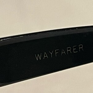 May include: Close-up of a black Wayfarer style eyeglass frame. The word "WAYFARER" is etched in white on the inner arm of the glasses. The frame is glossy and reflects the light.