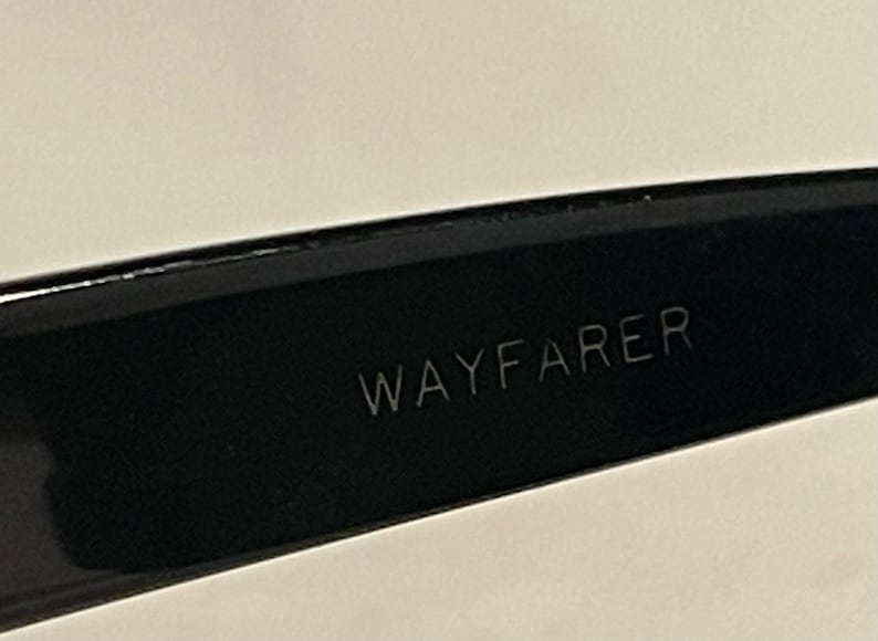 May include: Close-up of a black Wayfarer style eyeglass frame. The word "WAYFARER" is etched in white on the inner arm of the glasses. The frame is glossy and reflects the light.
