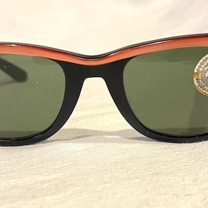 May include: Black-framed sunglasses with a coral-coloured upper rim and green lenses. The sunglasses have a classic design with a small, round emblem on the right lens. The lenses are slightly curved.