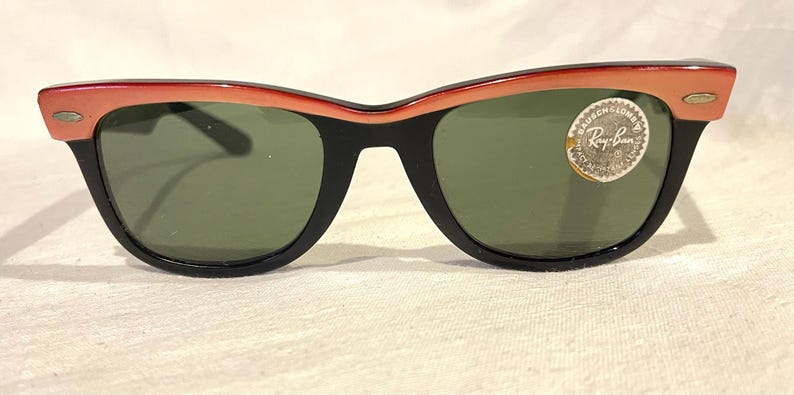 May include: Black-framed sunglasses with a coral-coloured upper rim and green lenses. The sunglasses have a classic design with a small, round emblem on the right lens. The lenses are slightly curved.