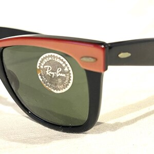 May include: Black and red sunglasses with green lenses. The sunglasses feature a classic design with a red upper frame and black arms. A circular sticker with text is on the lens.
