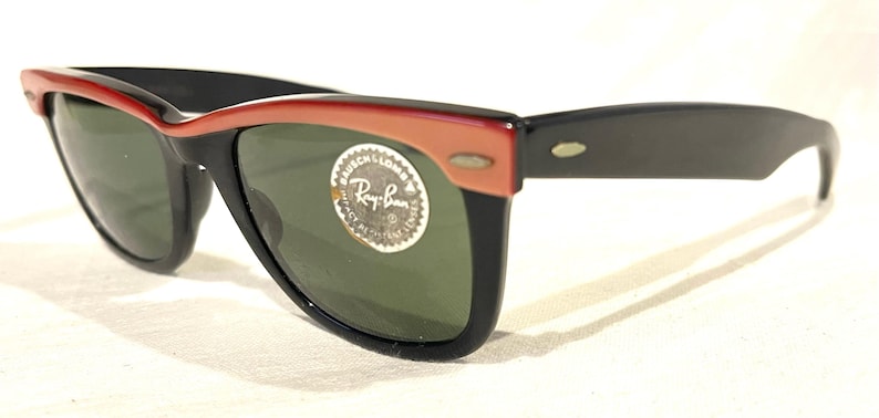 May include: Black and red sunglasses with green lenses. The sunglasses feature a classic design with a red upper frame and black arms. A circular sticker with text is on the lens.