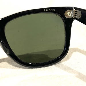 May include: Close-up of black sunglasses with green lenses. The frame is thick and glossy, with the text "B&L 5022" printed on the upper left corner. The hinge is visible, and the lenses are slightly reflective.