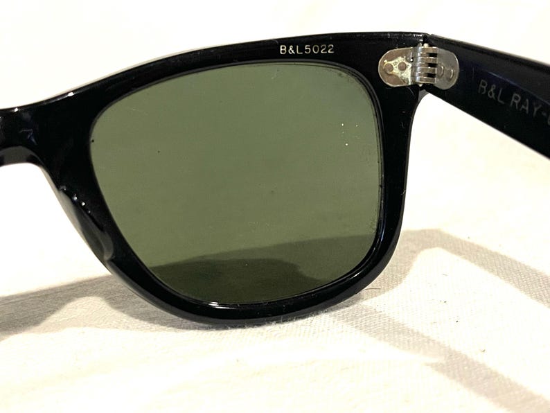 May include: Close-up of black sunglasses with green lenses. The frame is thick and glossy, with the text "B&L 5022" printed on the upper left corner. The hinge is visible, and the lenses are slightly reflective.