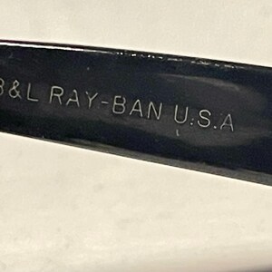 May include: Close-up of a black Ray-Ban sunglasses arm with the text "B&L RAY-BAN U.S.A" in silver lettering. The hinge is visible, with a silver metal finish. The sunglasses arm is glossy and reflects light.