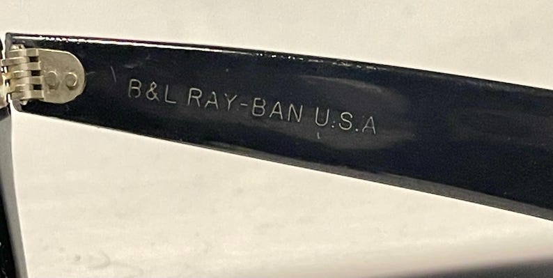 May include: Close-up of a black Ray-Ban sunglasses arm with the text "B&L RAY-BAN U.S.A" in silver lettering. The hinge is visible, with a silver metal finish. The sunglasses arm is glossy and reflects light.