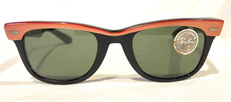 May include: Vintage Ray-Ban sunglasses. The black frame supports green lenses, with a red and peach gradient across the top. A round sticker with the Ray-Ban logo is on the right lens.