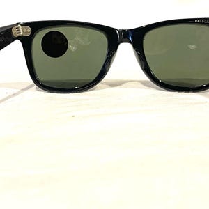 May include: Black sunglasses with green lenses, viewed from the inside. The frames are glossy and the arms extend outwards. The lenses are a muted green colour. The sunglasses are set against a white background.