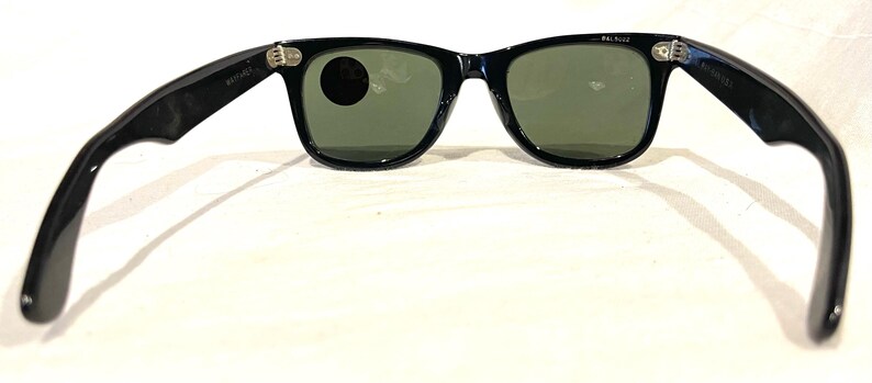 May include: Black sunglasses with green lenses, viewed from the inside. The frames are glossy and the arms extend outwards. The lenses are a muted green colour. The sunglasses are set against a white background.