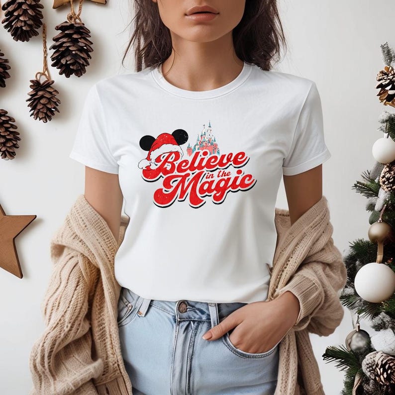 Peut inclure: T-shirt blanc avec un motif rouge et blanc de Mickey Mouse portant un bonnet de P&egrave;re No&euml;l et le texte "Believe in the Magic" avec un ch&acirc;teau en arri&egrave;re-plan.