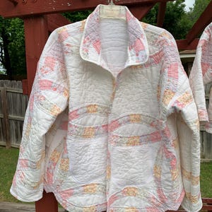 May include: A white quilted jacket with pink and yellow floral patchwork. The jacket features a collar and two front pockets. It is hanging on a hanger. The jacket is made from a soft, quilted fabric.