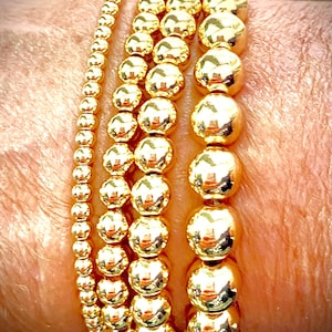 Może przedstawiać: Prezentowane są cztery złote bransoletki z koralików o różnych rozmiarach. Bransoletki wykonane są z błyszczących, okrągłych koralików, odbijających światło. Bransoletki są noszone na nadgarstku, prezentując różne rozmiary koralików i ogólny złoty kolor.