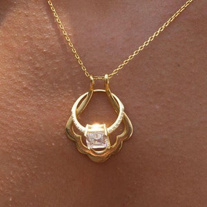 May include: A gold chain necklace with a pendant featuring a clear square-shaped gemstone set in a gold frame. The pendant is surrounded by a decorative gold design.