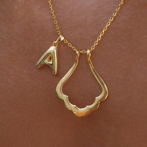 May include: A gold chain necklace with a small letter 'A' charm and a larger, open, ornate charm. The necklace is worn on a person's chest.
