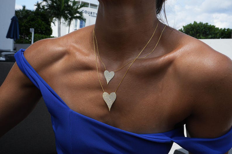 May include: A woman wearing a blue off-the-shoulder top with a gold chain necklace featuring two diamond-encrusted heart pendants.