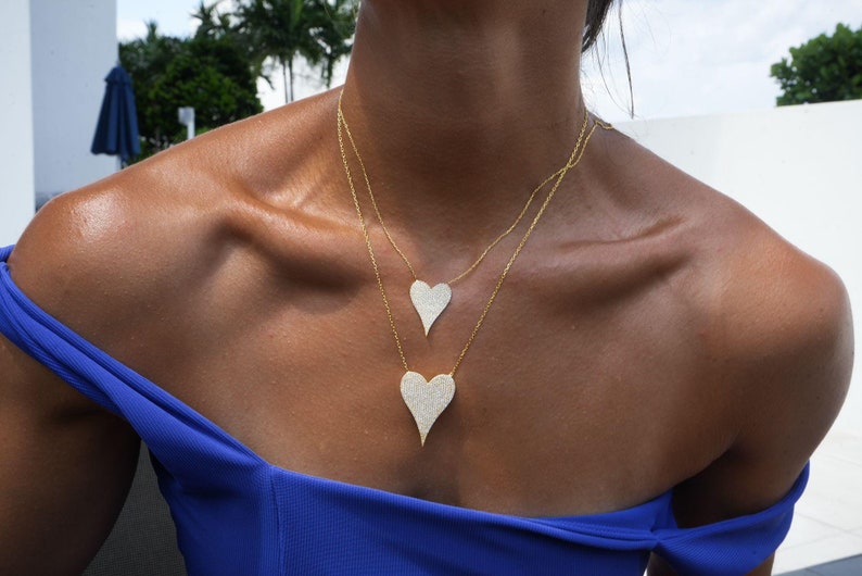 May include: A woman wearing a blue top with two gold necklaces featuring a heart-shaped pendant with a diamond-like design.