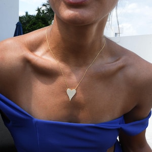May include: A gold necklace with a large, heart-shaped pendant. The pendant is made of a textured metal and has a shiny finish. The necklace is worn by a person with a tan complexion and is wearing a blue off-the-shoulder top.