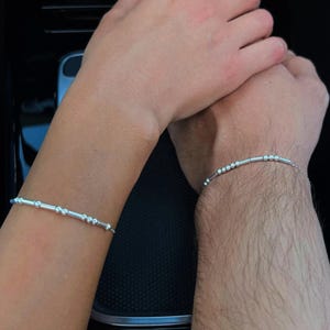 May include: Two silver bracelets with small round beads and a bar design. The bracelets are on the wrists of two people.