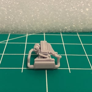 Puede incluir: Un pequeño modelo gris de un motor, con componentes detallados como un turbocompresor y tubos de escape. El modelo se encuentra sobre un fondo de cuadrícula verde, sugiriendo una escala o contexto de medición.